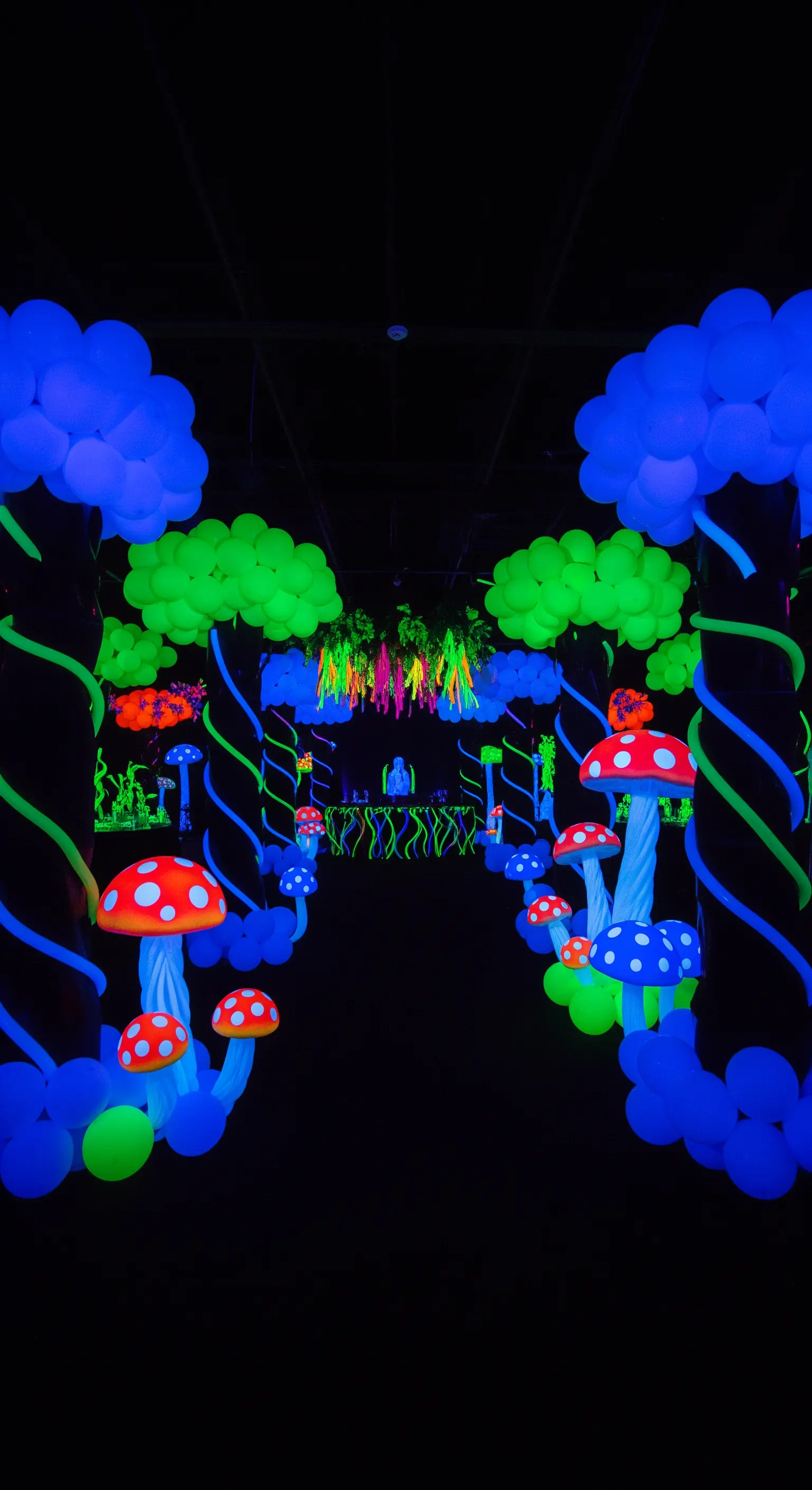 Corridoio buio decorato con palloncini fluorescenti a forma di funghi e alberi sotto luce UV.