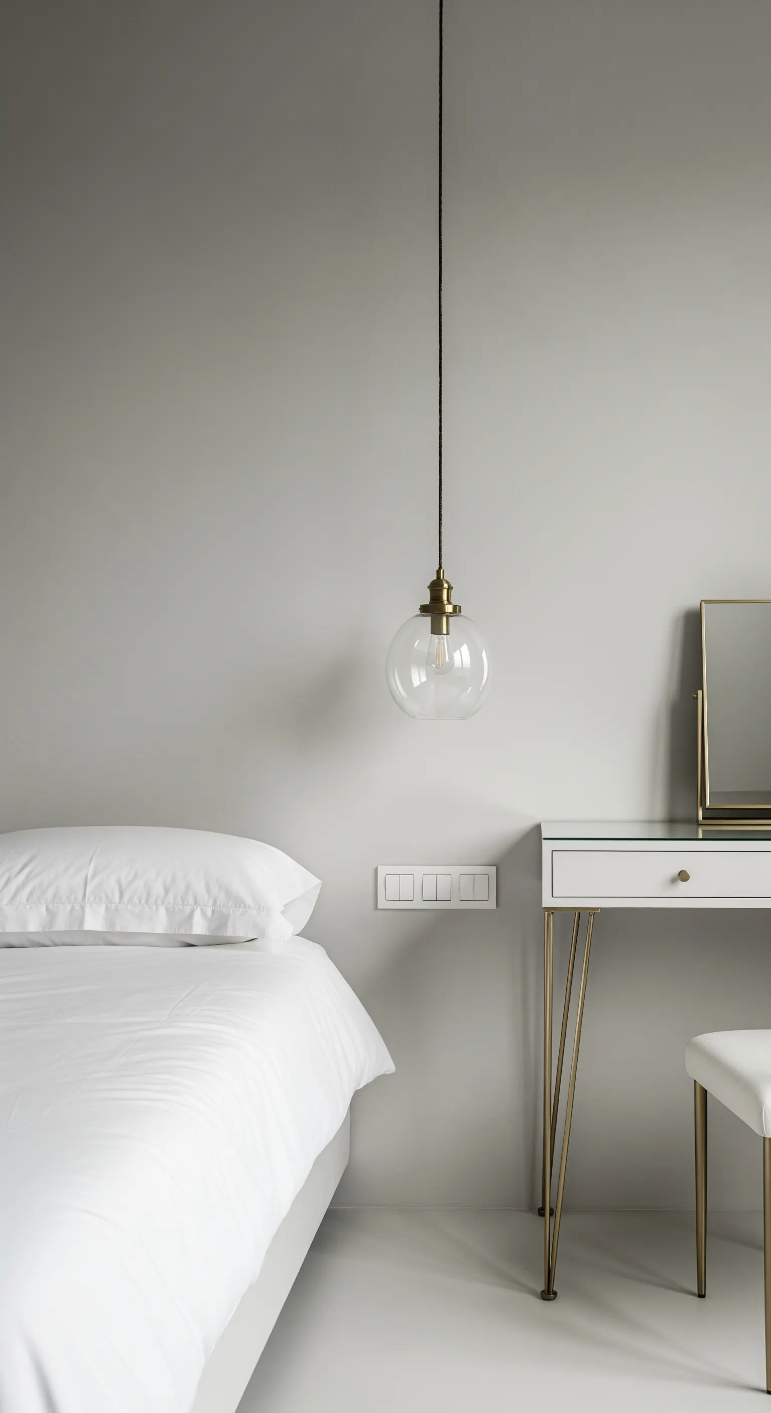 Lampada a sospensione in vetro e ottone appesa accanto a un letto con parete grigio chiaro.