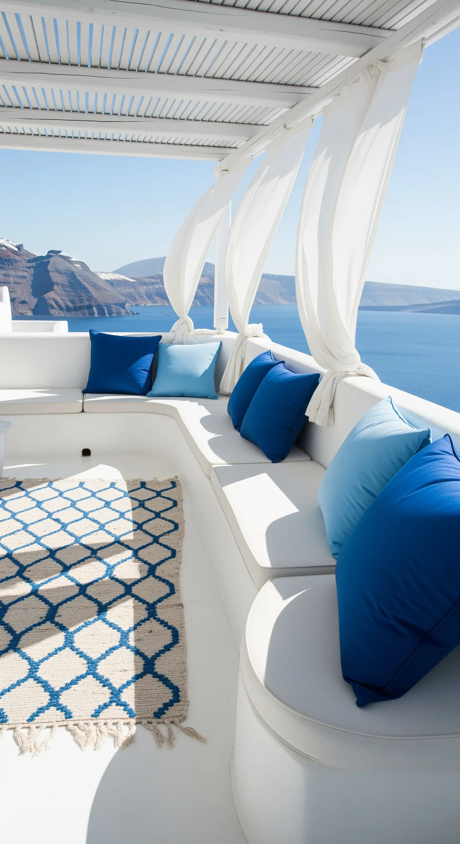 Terrazza bianca a Santorini con sedute in muratura, cuscini blu e vista sul mare.