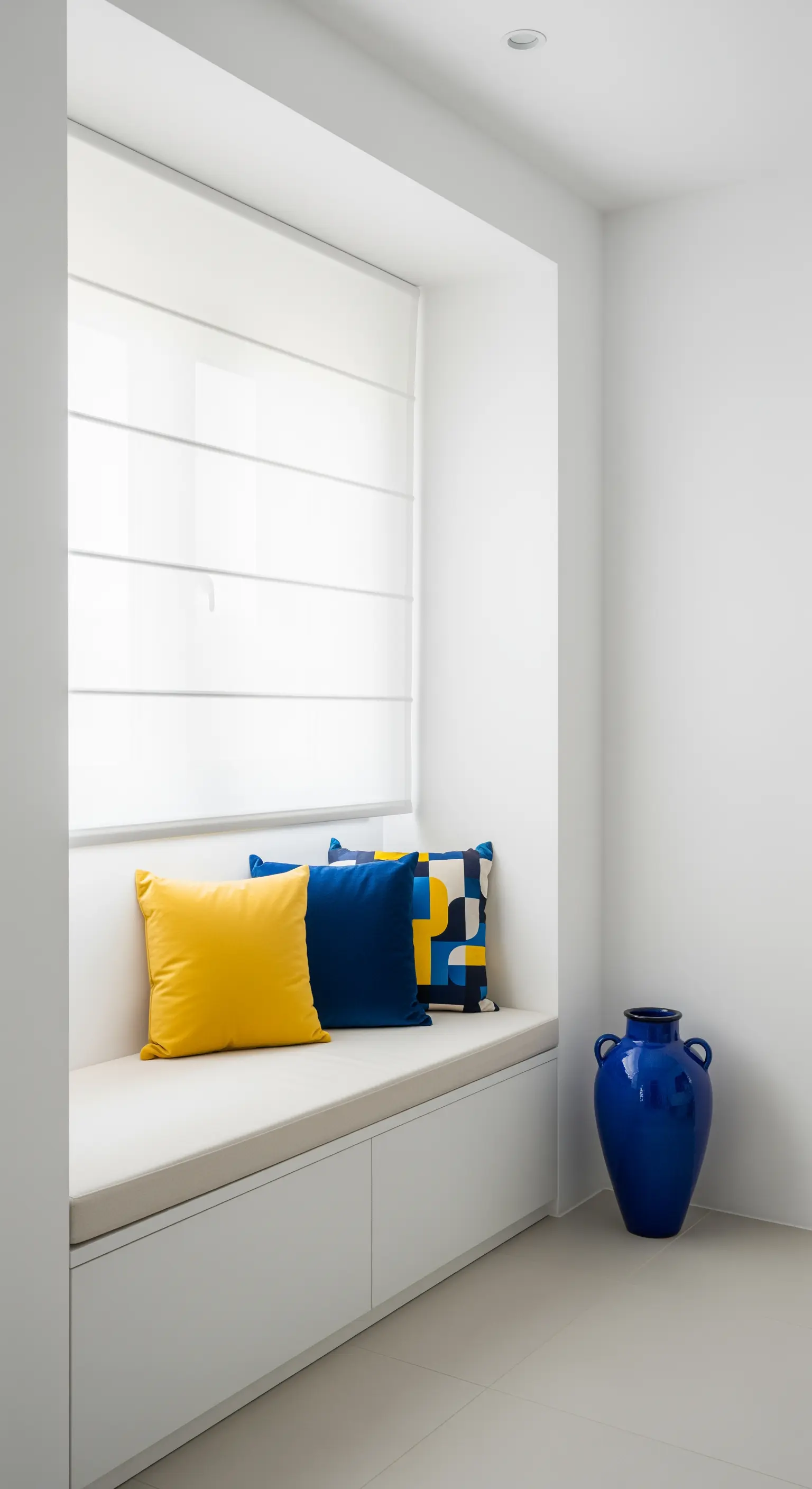 Nicchia bianca e minimale con panca, cuscini gialli e blu e un grande vaso blu a terra.