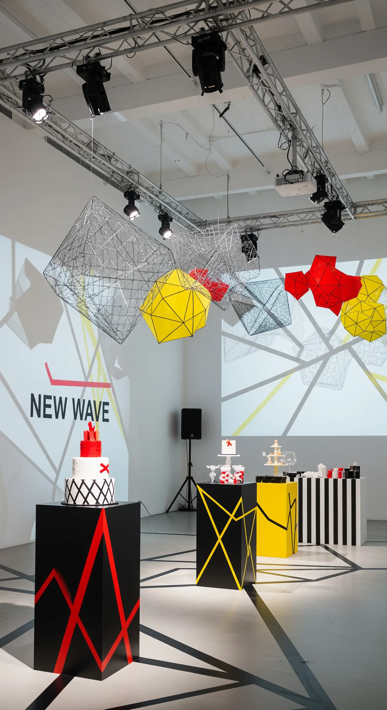 Allestimento in stile New Wave con piedistalli neri, grafiche geometriche e installazioni sospese.