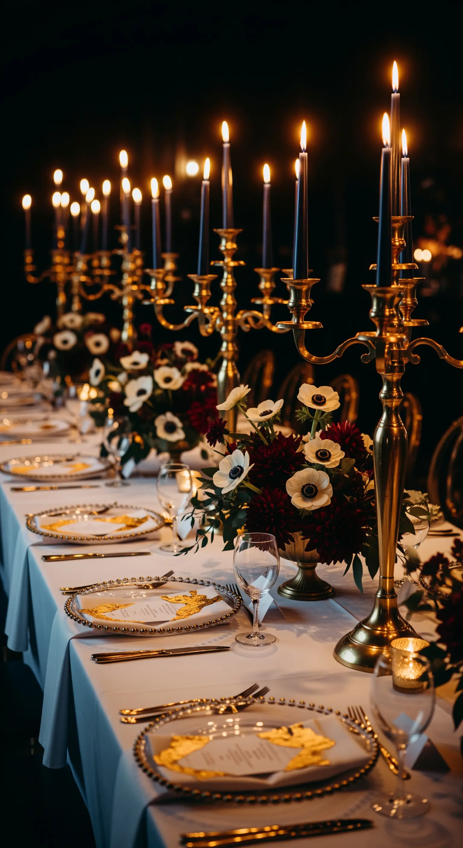 Tavola elegante al buio con candelabri dorati, candele nere e fiori bianchi e bordeaux.