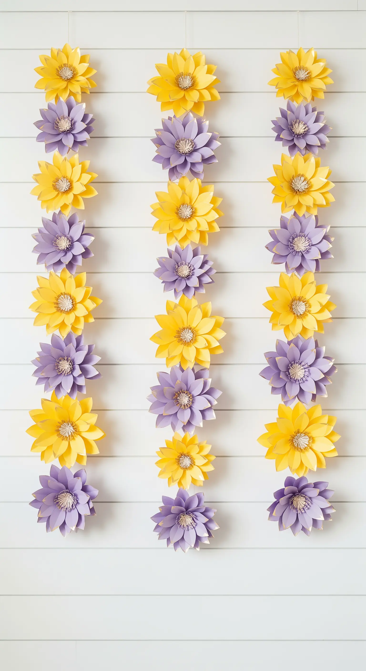 Tre ghirlande verticali di fiori di carta gialli e viola appese a una parete bianca a listelli.