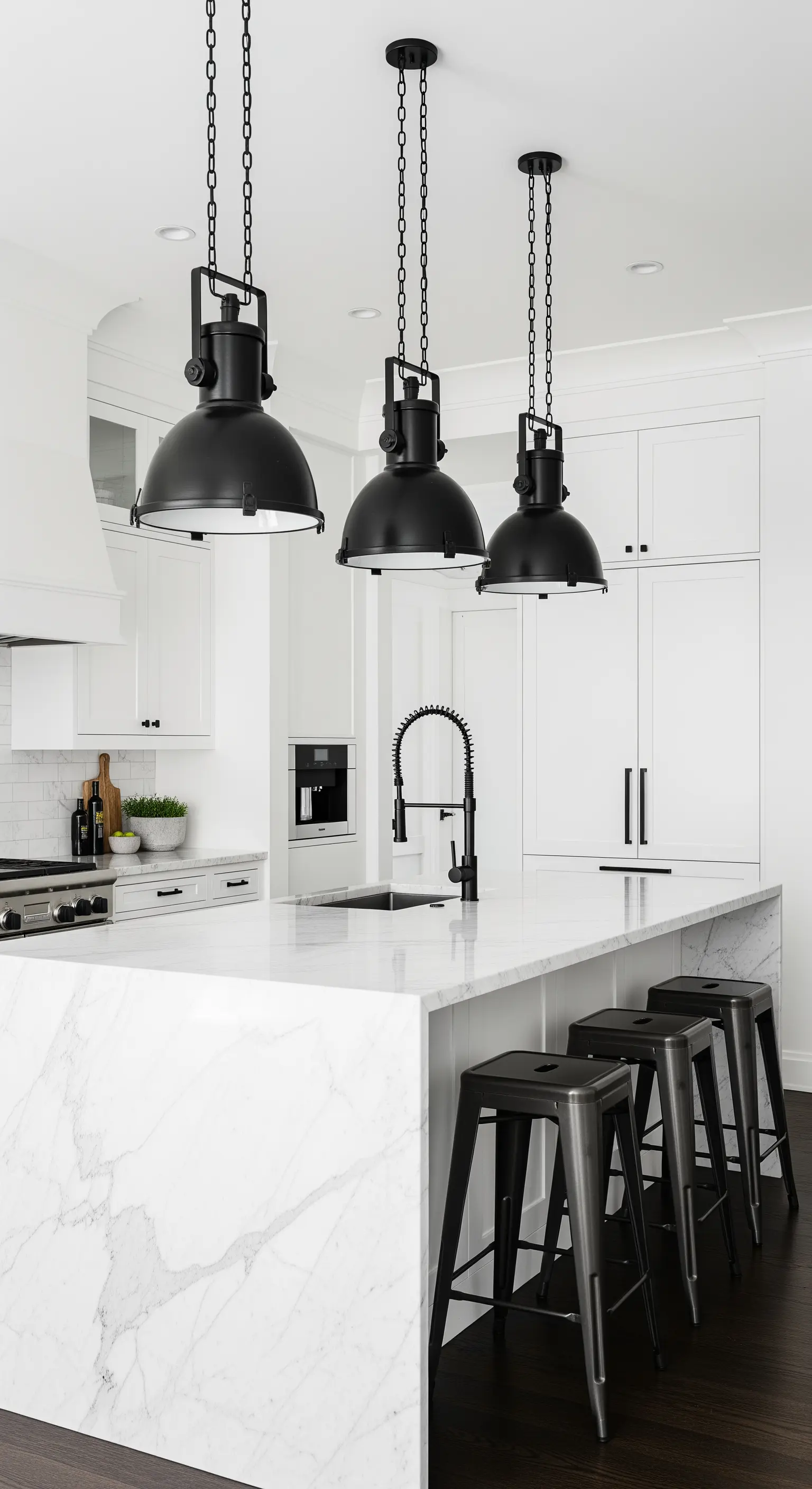 Cucina total white con isola in marmo, lampade industriali e sgabelli neri a contrasto.