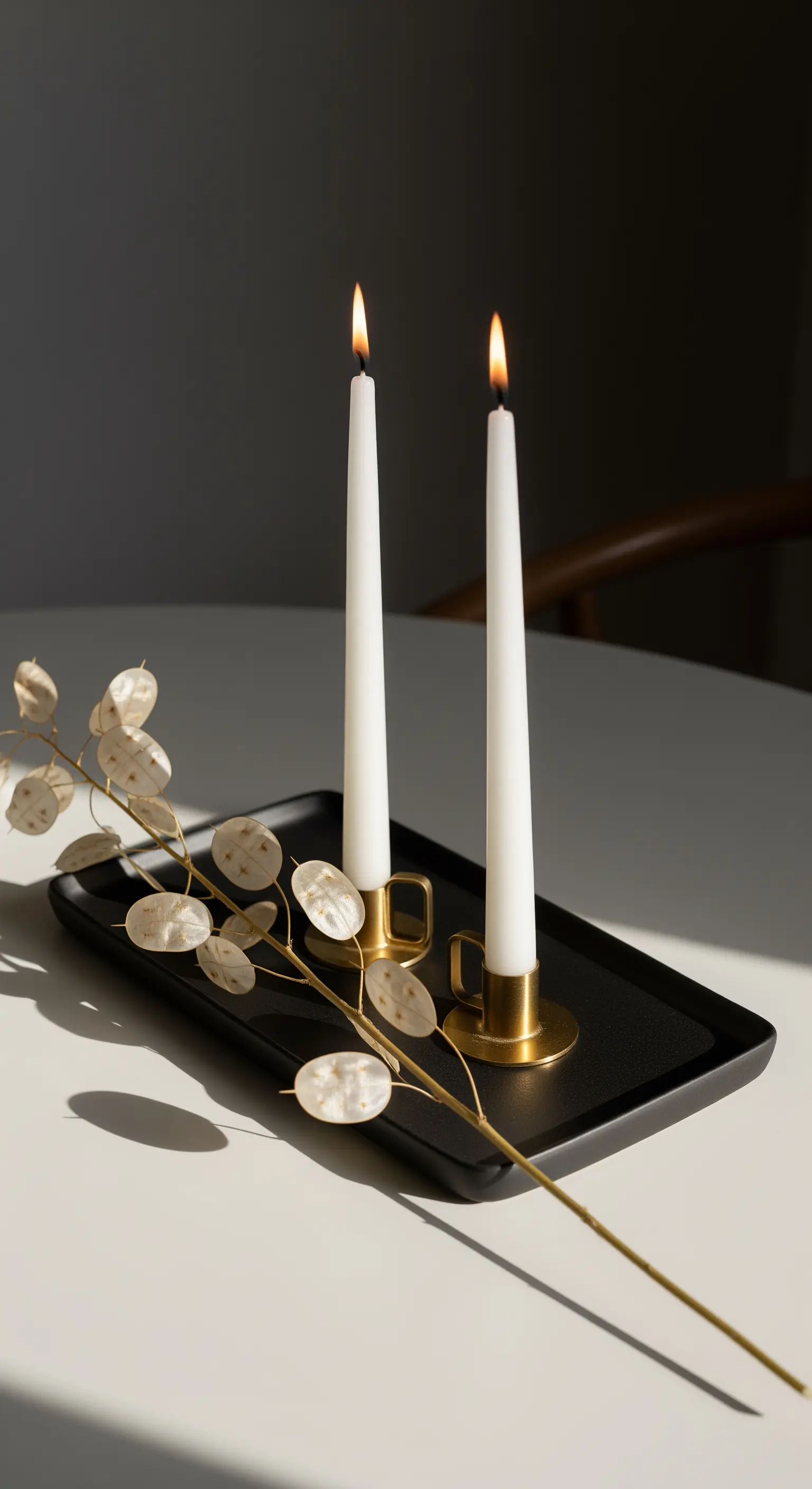 Vassoio nero minimalista con candele bianche in portacandele dorati e ramo di lunaria.