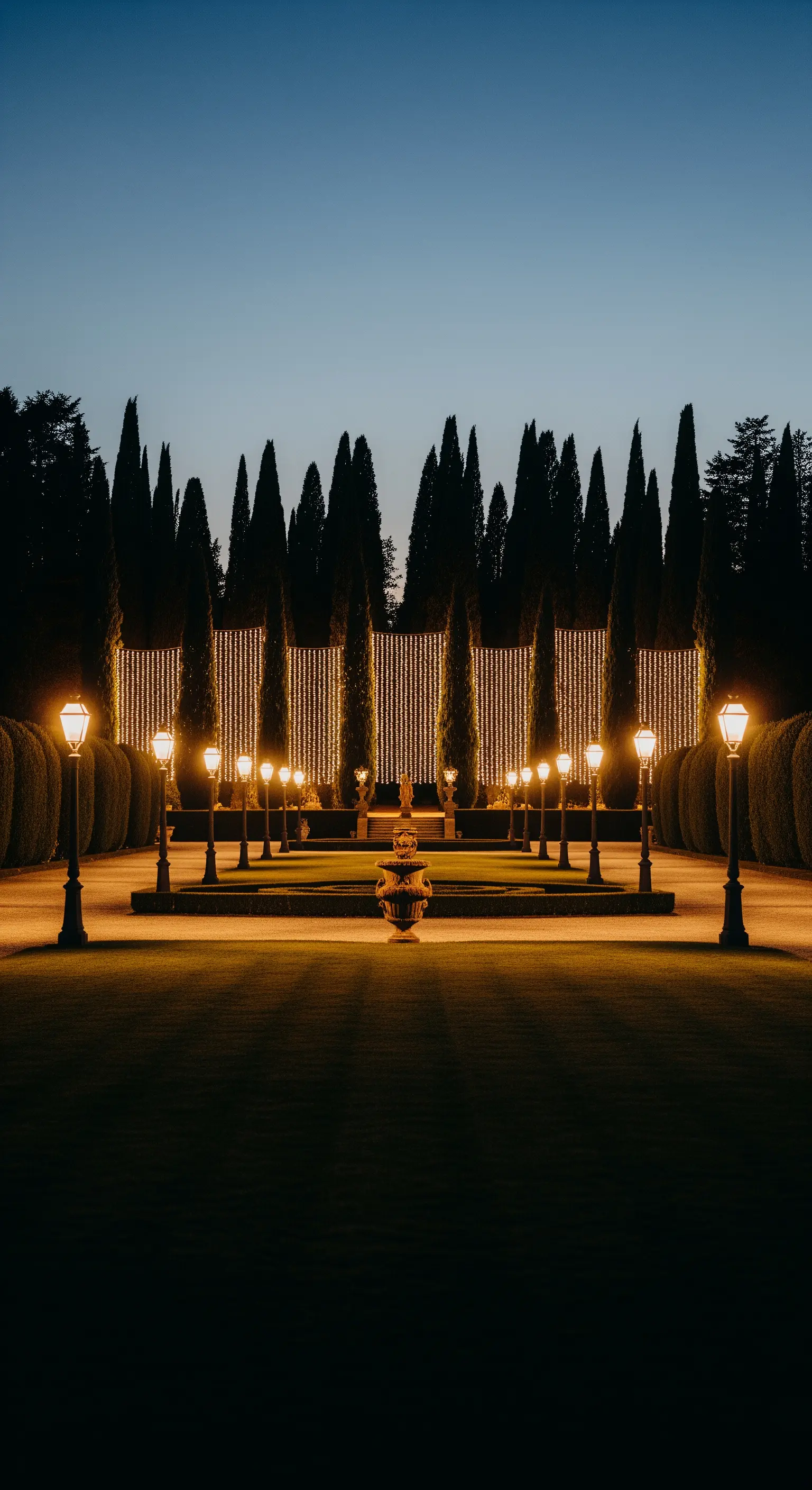 Giardino formale con cipressi, lampioni simmetrici e una parete di luci a cascata sullo sfondo.