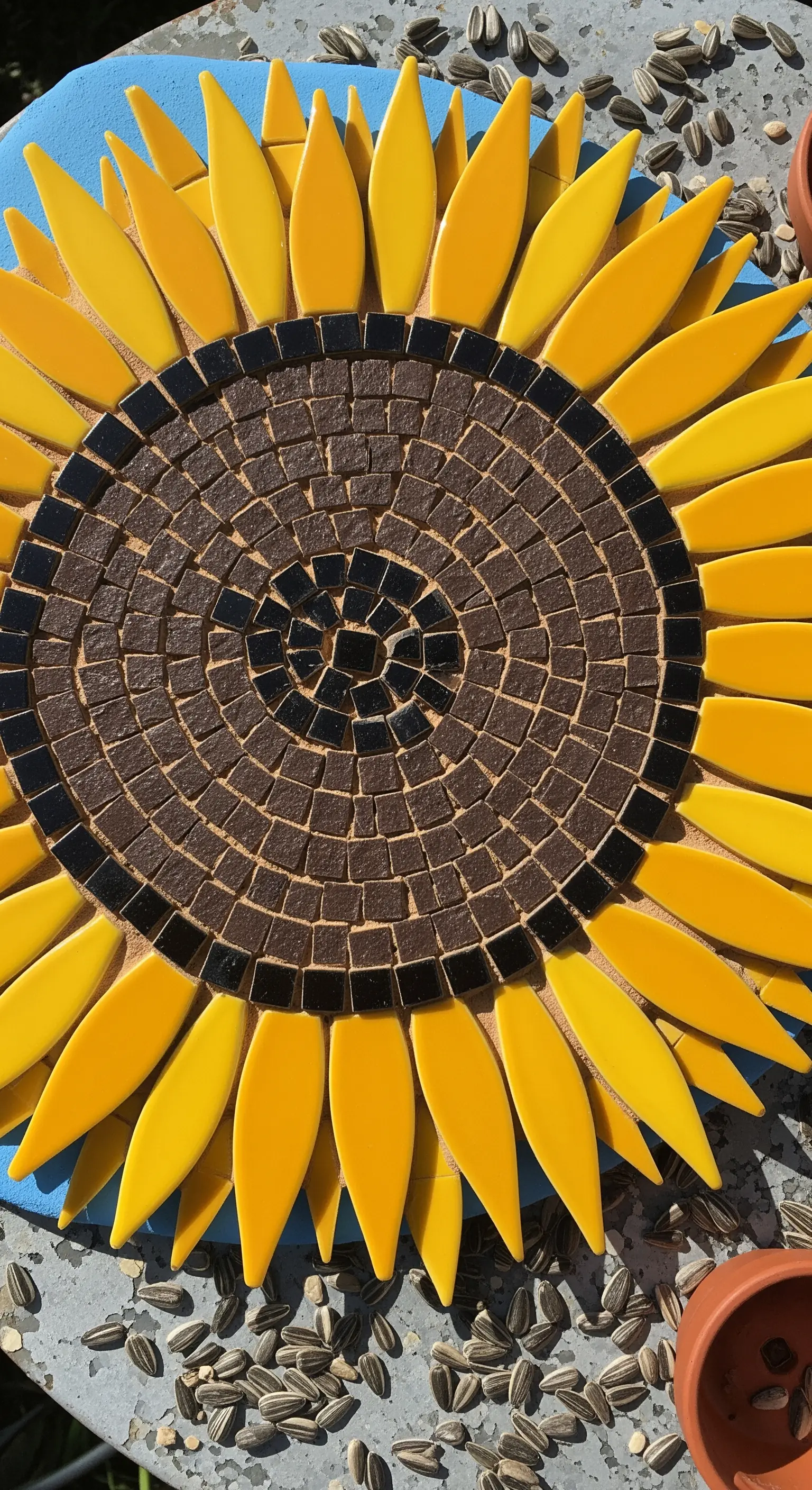 Dettaglio di un mosaico a forma di girasole, con il centro a spirale e i petali gialli allungati.