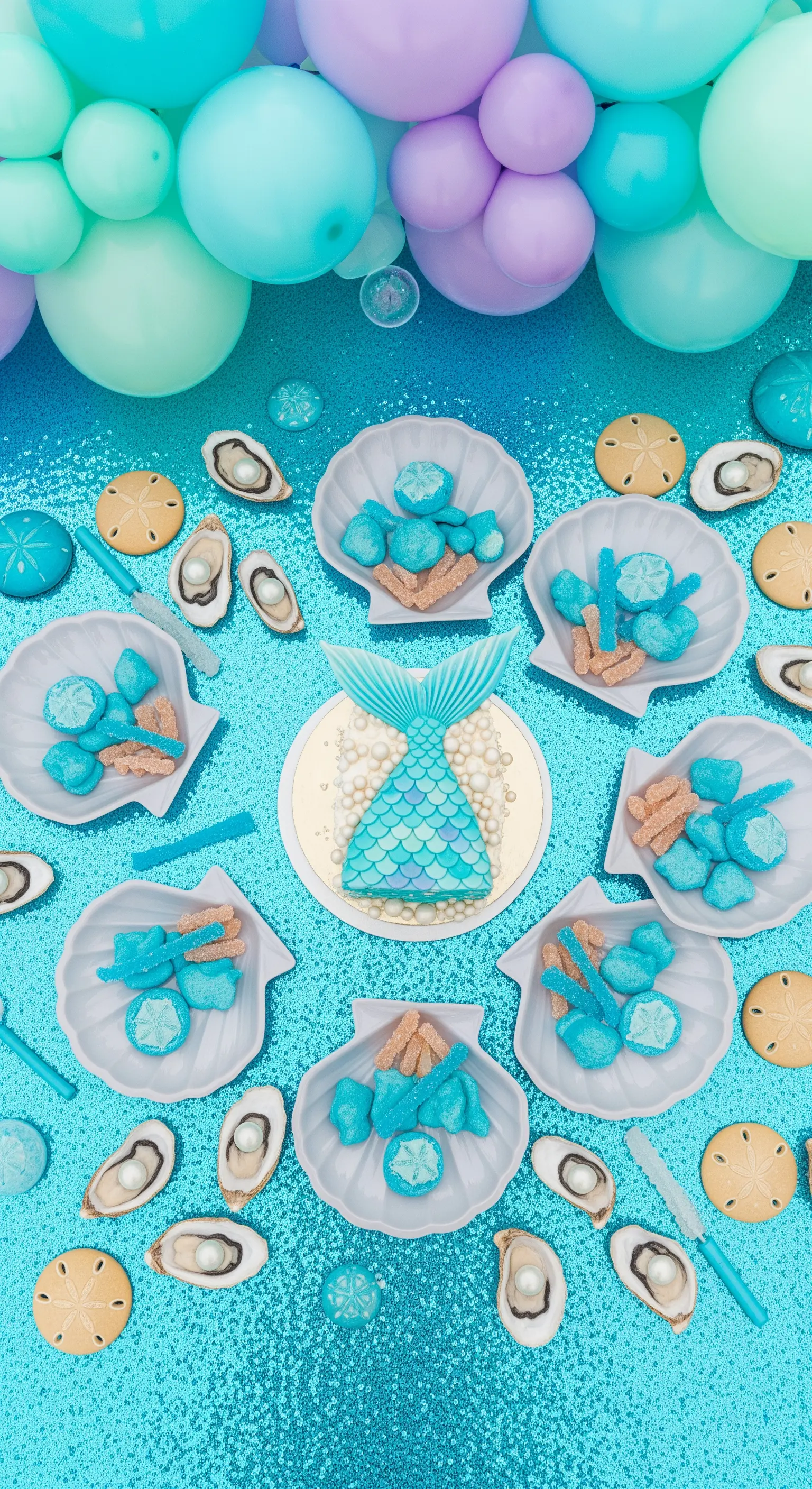 Allestimento a tema sirena con tovaglia di paillettes, dolci in conchiglie e coda di sirena.