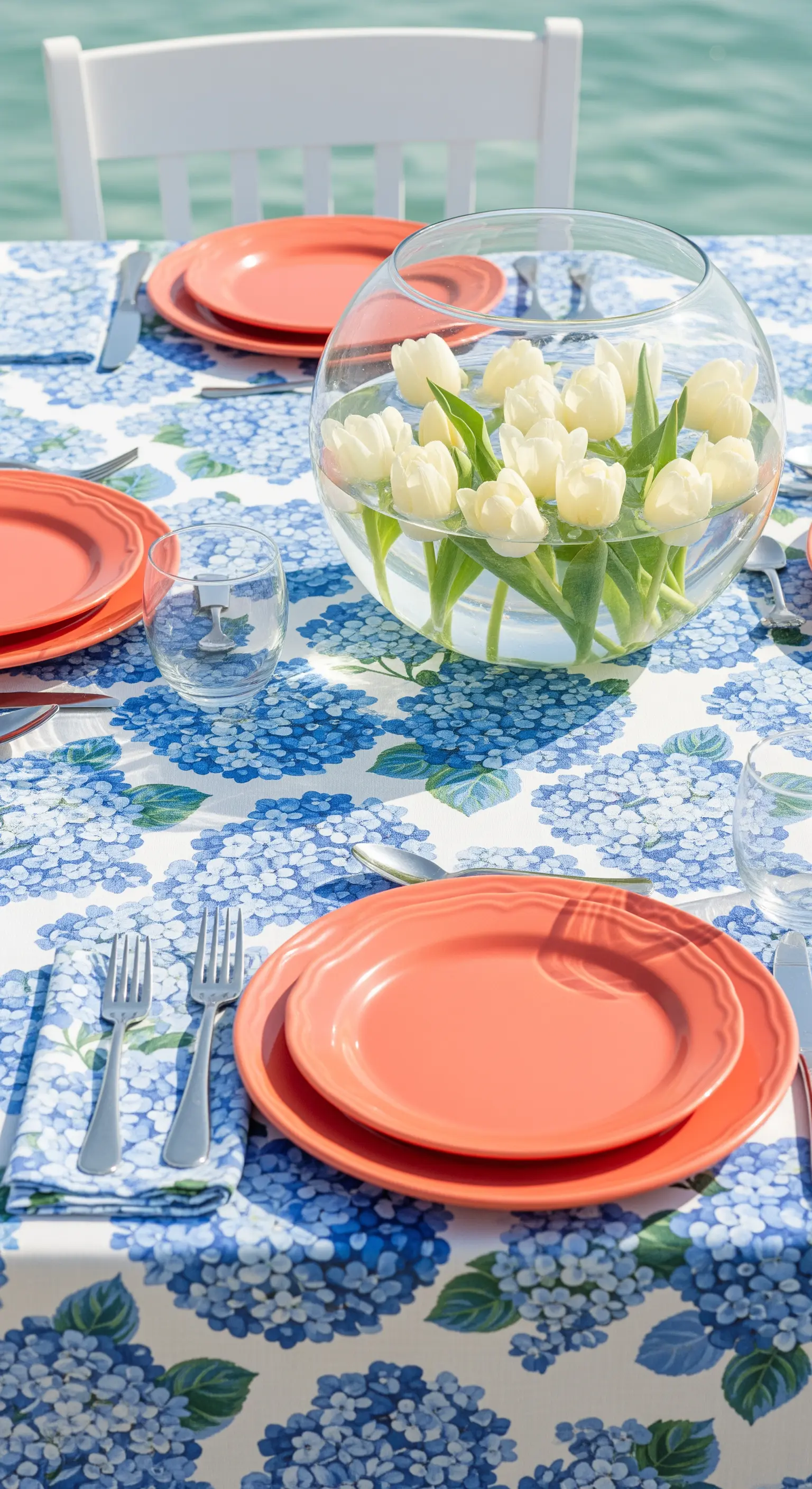 Tavola con tovaglia a ortensie blu, piatti corallo e tulipani bianchi in una boccia d'acqua.