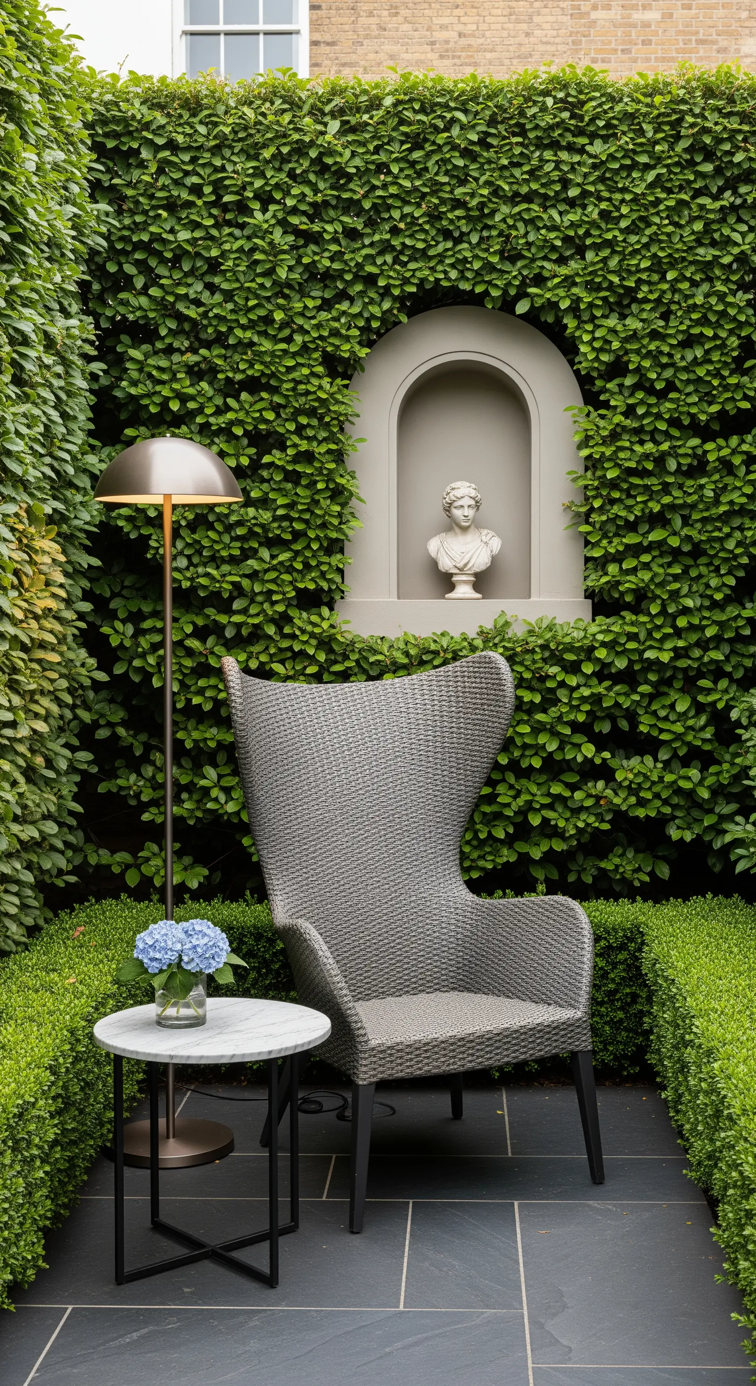 Angolo lettura in giardino con poltrona in vimini, tavolino in marmo e un busto in una nicchia